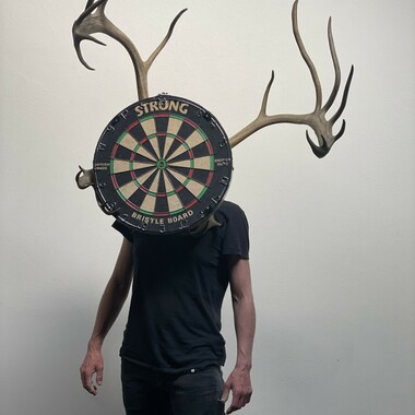 Ein Körper von der Taille aufwärts. Vor dem Gesicht ist eine Dart-Scheibe mit einem Geweih.