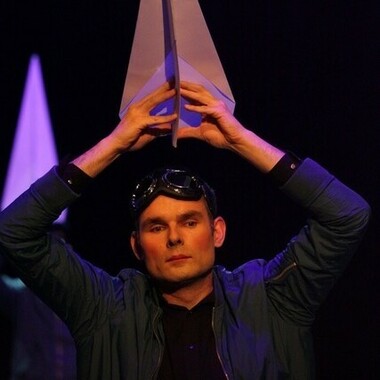 Ein*e Performer*in in einem Pilot*innen-Kostüm hält sich einen großen Papierflieger über den Kopf.