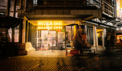 Pictured is the Ballhof Zwei building at night while people are walking by. Above the entrance is a glowing sign in yellow lettering reading Ballhof Zwei.