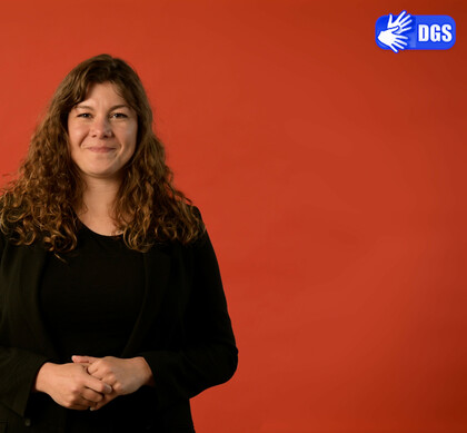 Athina Lange vor einem einfarbigen orangefarbenen Hintergrund. Oben rechts im Bild ein blaues DGS Piktogramm mit zwei gebärdenden Händen.