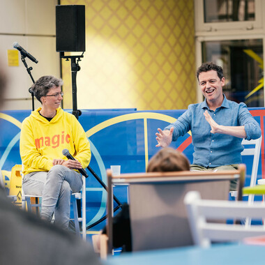 Anna Mülter und Richard Dougherty im Gespräch. Sie sitzen im Festivalzentrum. Richard gebärdet BSL, Anna schaut ihn aufmerksam an.