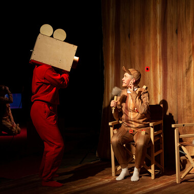 A stage covered in a brown fabric with a wooden look. On the right are two wooden chairs as they are on a film set. In the left chair sits Ivor MacAskill. Ivor is wearing a brown costume consisting of baseball cap, sweater and pants with a wooden look and white socks. In his right hand, Ivor holds a microphone with a plush headpiece and looks up at the person standing in front of him on the left. The person is wearing a loose tracksuit in a rich red tone. The face is not visible, because the person has a large wooden box on their shoulder, which has the shape of a camera.