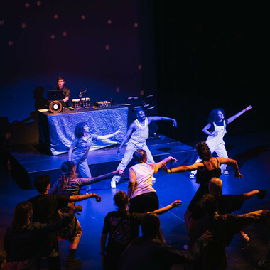 SPIN in der Cumberlandschen Bühne: Anna Seymour (links), Chris Fonseca (Mitte) und Raffie Julien (rechts) tanzen in einem Raum, der in blaues Licht eingetaucht ist. Das Publikum vor ihnen ahmt ihre Armbewegungen nach. Hinter Anna, Chris und Raffie ist eine DJ-Pult zu sehen, hinter dem eine Person sitzt, die ein Laptop bedient. Auf dem Pult befindet sich unter anderem eine Bongo-Trommel.