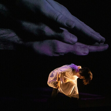 Chisato Minamimura in a white knee-length robe. She kneels on the stage with her back bent forward, looking down at the floor. At the same time, her right hand rests on one thigh. Three oversized hands are projected onto the dark wall behind her, lying flat on top of each other. Compared to the projection, Chisato appears very small.