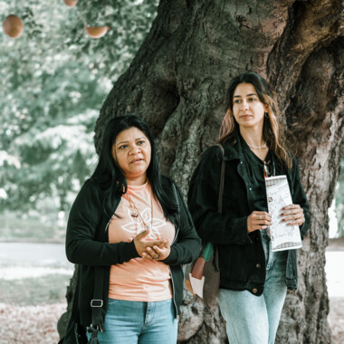 Two people stand next to each other in front of a tree and seem to be talking.