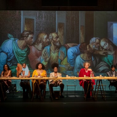 Eine Szene aus dem Stück “The Last Supper” von MEXA. Es zeigt sechs Personen an einem langen Tisch auf der Bühne zu sitzen. Auf dem Tisch sind Teller und Becher zu sehen. Hinter ihnen eine Wand, auf die ein Teil des Gemäldes “Das Abendmahl” projiziert wird.