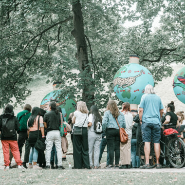 A group of about 20 people stand in front of itaakás hanging in the trees of the theatre park.
