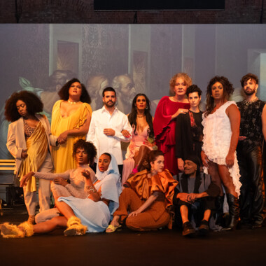 A scene from the play ‘The Last Supper’ by MEXA. This is a group photo of the MEXA collective. Everyone is festively dressed and posing on stage. In the front row, four members of the collective sit next to each other on the floor. Behind them, eight MEXA members can be seen standing next to eachother. Behind them is a wall onto which part of the painting ‘The Last Supper’ is projected.