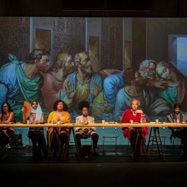 A scene from the play ‘The Last Supper’ by MEXA. It shows six people sitting at a long table on the stage. Plates and cups can be seen on the table. Behind them is a wall onto which part of the painting ‘The Last Supper’ is projected.