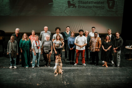 Gruppenfoto: Alle Redner*innen der Veranstaltung “Wir verbünden uns” stehen als Gruppe zusammen, vor ihnen sitzt ein Hund und schaut die mittig stehende Person an.