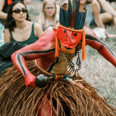 Ziel Karapotó während der Performance Confluences. Er trägt eine Maske die an einen Fisch erinnert, einen Kopfschmuck aus blauen Federn und einen Baskrock. Der Oberkörper rot geschminkt, mit zwei schwarzen Streifen die sich jeweils vom Handgelenk bis zur Hüfte ziehen. In der Hand hält er eine Rassel.