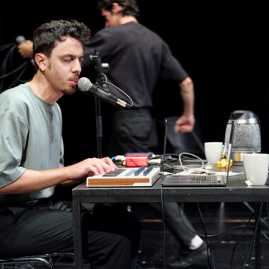Ein Mensch sitzt auf der Bühne an einem Tisch mit Laptop, MIDI-Keyboard und Mikrofon und singt mit geschlossenen Augen. Im Hintergrund bewegen sich zwei weitere Personen, teils unscharf. Auf dem Tisch stehen weiße Tassen und eine Thermoskanne.