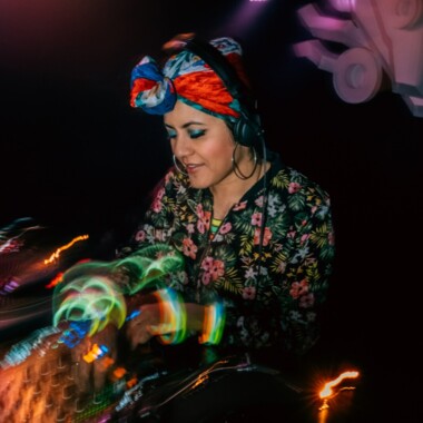 DJ mit buntem Kopftuch und Kopfhörern legt Musik auf. Im Vordergrund sind bunte verschwommene Lichter. 