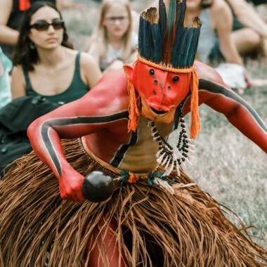 Ziel Karapotó during the performance Confluences. He wears a mask reminiscent of a fish, a headdress made of blue feathers and a basque skirt. His upper body is painted red, with two black stripes running from his wrists to his hips. He holds a rattle in his hand.