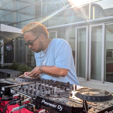 Yoel mit Sonnenbrille spielt draußen an einem Pioneer-Mischpult, im Hintergrund ist ein Gebäude mit großen Fenstern, Sonnenlicht fällt von hinten ein.