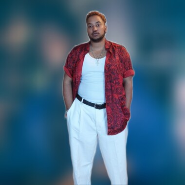 Yoel trägt ein weißes Top, eine weiße Hose und ein rotes gemustertes Hemd, der Hintergrund ist unscharf. 