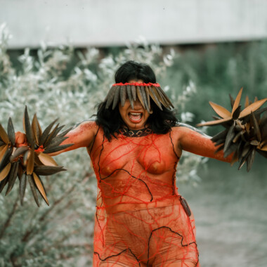Lilly Baniwa währen der Performance Confluences. Sie ist am ganzen Körper orangefarben geschminkt und trägt einen Kopfschmuck der ihrer Augen verdeckt. Ihr Gesicht ist ausdrucksstark, sie hat den Mund geöffnet als würde sie etwas rufen. In den Händen hält sie Rasseln.