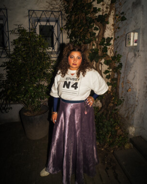 Person mit weißen Stussy-Pullover und lila Rock, steht in einem Hof bei Nacht, im Hintergrund Gebäude und Pflanzen.