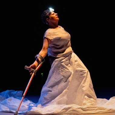 A scene from the performance ‘The Songs of the Wayfarer’ by Claire Cunningham: It shows Claire Cunningham with crutches on a dark stage. Claire has short grey hair and is wearing a long white dress and a headlamp.