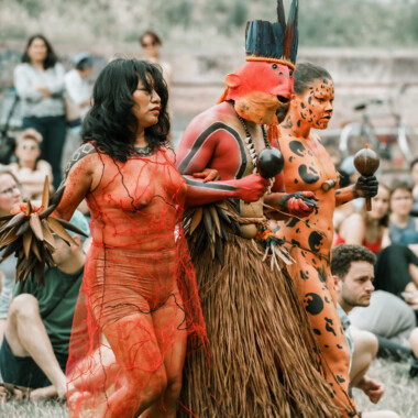 A scene from the performance Confluences. Lilly Baniwa, Ziel Karapotó, and Olinda Tupinambá stand close together, all holding rattles in their hands. They are all wearing striking costumes. One person is dressed as a jaguar, another wears a mask reminiscent of a fish, and another is painted orange all over their body. In the background, the audience sits on the lawn.