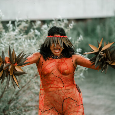Lilly Baniwa during the performance Confluences. Her entire body is painted orange and she wears a headdress that covers her eyes. Her face is expressive, her mouth open as if she were shouting something. She holds rattles in her hands.