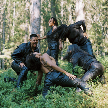 A scene from the work  ‘Undersang’ by Harald Beharie: Seven people, all dressed in black leather, are standing in a forest. The person on the far left is standing and sticking out their tongue, three people in the centre of the picture are crouching and kneeling on the forest floor and appear to be screaming. Two people on the right are standing and appear to be moving.