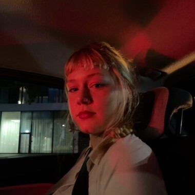 Eine Person mit blondem Haar sitzt in einem Auto, sie blickt in die Kamera, ihr Gesicht wird von rotem Licht beleuchtet.