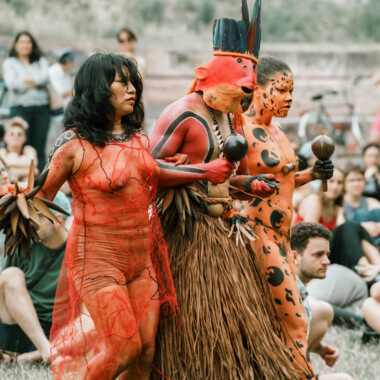 Eine Szene aus der Performance Confluences. Lilly Baniwa, Ziel Karapotó, Olinda Tupinambá stehen eng beieinander, alle haben Rasseln in den Händen. Sie tragen alle ein auffälliges Kostüm. Eine Person ist als Jaguar geschminkt, eine trägt eine Maske die an einen Fisch erinnert, eine ist über den gesamten Körper orangefarben geschminkt. Im Hintergrund befindet sich das Publikum, das auf der Wiese sitzt.