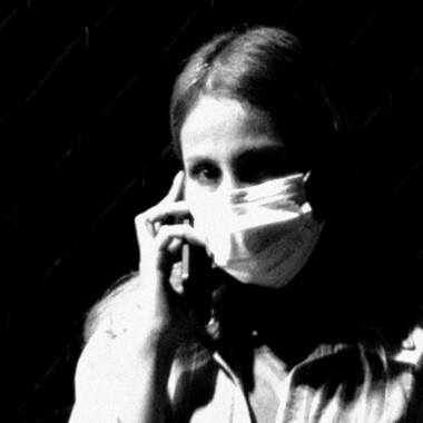 Ein schwarz/weiß Foto. Eine Person mit langen Haaren und weißer OP-Maske hält ein Handy ans Ohr. Der Blick ist direkt in die Kamera gerichtet. Im Hintergrund ist ein Gitter zu sehen.