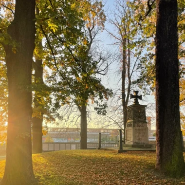 Eine parkähnliche Anlage mit hohen Bäumen im Herbst. Das Sonnenlicht fällt durch die Bäume und Blätter, die größtenteils bereits am Boden liegen. Auf einem kleinen Hügel steht ein steinernes Denkmal mit einem Kreuz oben drauf.