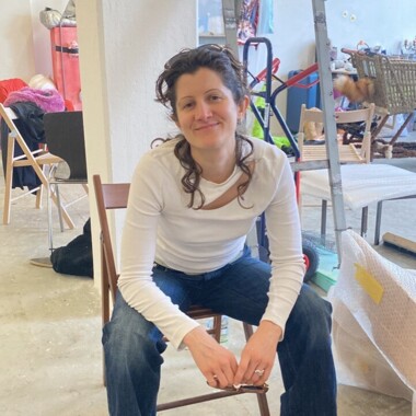 Eine Person sitzt in einem Atelier oder Proberaum auf einem Holzstuhl und blickt lächelnd in die Kamera. Sie trägt Jeans und ein weißes Shirt. Im Hintergrund stehen weitere Stühle, Tische und Materialien.