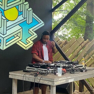 Eine Person steht hinter einem DJ-Setup auf einer überdachten Bühne und trägt Kopfhörer. Hinter dem Pult befinden sich eine großformatige Grafik und Bäume, auf dem Tisch stehen ein Mischpult und Audiotechnik.