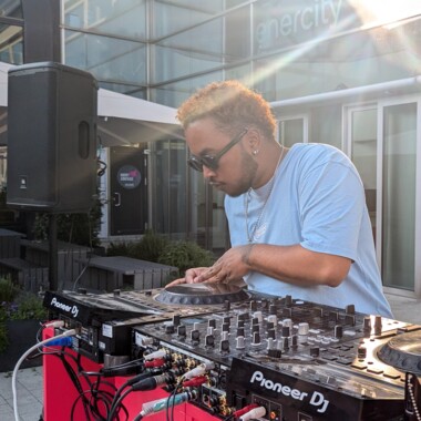Eine Person mit Sonnenbrille und hellblauem T-Shirt bedient ein DJ-Pult im Freien. Im Vordergrund sind Mischpult und Plattenspieler, im Hintergrund moderne Gebäude mit vielen Glasscheiben.