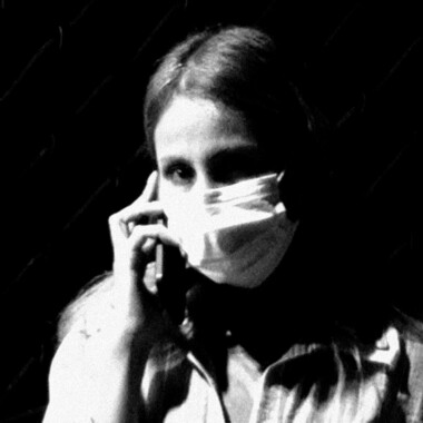 A black-and-white photograph. A person with long hair and a white surgical mask is holding a mobile phone to their ear. They are looking directly at the camera. A grille can be seen in the background.