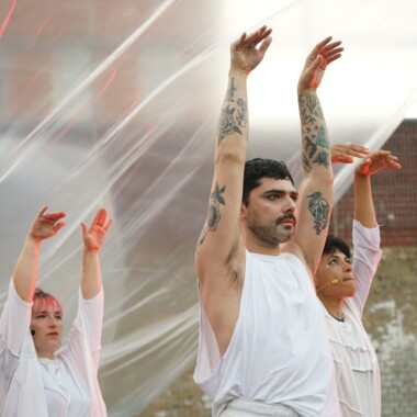 A performance in the courtyard of Rennelberg Prison. Three performers in white costumes raise their arms aloft. In the background are transparent sheets.
