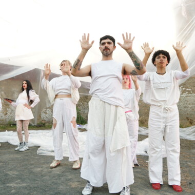 A performance in the courtyard of Rennelberg Prison.  Five performers in white costumes stand in front of an installation made of painter’s plastic sheeting, holding both hands upwards with their palms facing upwards. In the background are a brick wall and transparent sheets of plastic.