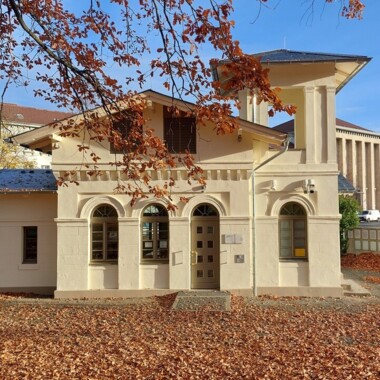 Die Gedenkstätte Schillstraße, ein helles, historisch wirkendes Gebäude. Es hat große Fenster, Säulen und einen klassischen Baustil. Der Boden davor ist mit Herbstlaub bedeckt, der Himmel im Hintergrund ist blau.