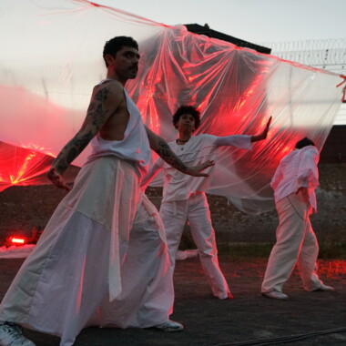 A performance in the courtyard of Rennelberg Prison. Three performers in white costumes move in front of a large-scale foil installation illuminated in red. They appear to be dancing with their arms outstretched.