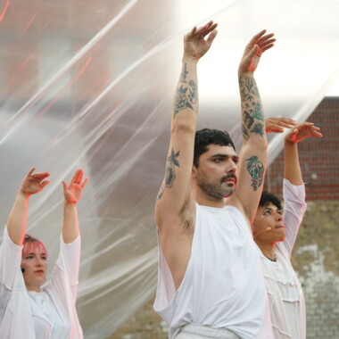 Eine Performance im Innenhof der JVA Rennelberg. Drei Performer:innen in weißen Kostümen heben die Arme in die Höhe. Im Hintergrund sind transparente Folien.