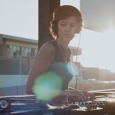 Eine Person mit Kopfhörern steht an einem DJ-Setup mit Plattenspielern und Mischpult auf einer Dachterrasse. Im Hintergrund sind Gebäude zu sehen, das Gegenlicht der Sonne beleuchtet die Szene.