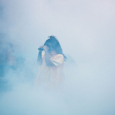 Eine Person in einem gelben Regenponcho steht in dichtem, bläulichem Nebel, nur ihr Oberkörper ist schemenhaft zu sehen. Sie hält ein Fernglas zwei Taschenlampen vor die Augen, als würde sie durch ein Fernglas blicken. Die Figur ist nur schemenhaft sichtbar, was eine geheimnisvolle, entrückte Atmosphäre erzeugt.