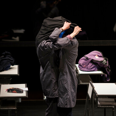 Zwei Schülerinnen in dunkler Schuluniform in der Bildmitte. Sie stehen eng umschlungen inmitten von weißen Schreibtischen. Sie bedecken ihre Köpfe gegenseitig mit einem schwarzen Tuch. Auf dem Schreibtisch hinter ihnen liegt ein lilafarbener Rucksack.