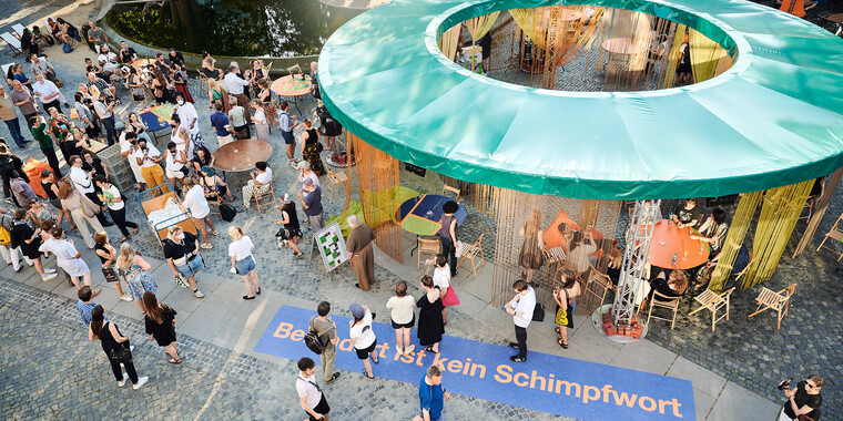 Bildbeschreibung seitens der Festival Theaterformen Redaktion. Selbstbeschreibung der abgebildeten Personen folgt. / Das Festivalzentrum auf dem Herzogin-Anna-Amalia-Platz von oben fotografiert. Auf der rechten Seite ist ein kreisrundes, überdachtes Pavillion mit dunkelgrünem Dach und Fädenvorhängen an allen Seiten. Überall stehen Menschen sowie mehrere runde Tische und Stühle verteilt. Unten im Bild ist eine dunkelblaue Bodenbeklebung mit der orangenen Aufschrift „Behindert ist kein Schimpfwort“ zu sehen. Oben links ist ein Teil des mit Wasser befüllten Brunnens zu sehen.