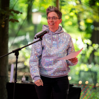 Anna Mülter during an opening speech. Anna has short grey hair, wears glasses and stands behind a microphone. She is holding a sheet of paper in her hand. She is wearing a grey, glittery hoodie and dark trousers. Behind her are trees.