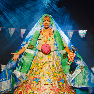 Eine Person auf einer Theaterbühne, die ein auffälliges, farbenfrohes Kostüm trägt. Das Kleid besteht aus verschiedenen Materialien, darunter bedruckte Stoffbahnen und Elemente, die an recycelte Plastiktüten erinnern. Der Rock ist mit bunten Blumenmotiven und einem großen, pinken Herz verziert. Die Person hält in einer Hand eine kleine Figur und trägt auffälligen Schmuck. 