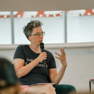 A person sits in a green armchair, speaking into a microphone and gesturing with their left hand. They wear glasses, a dark blue T-shirt with the words “THE FUTURE IS ACCESSIBLE,” and pink shorts. Blurred audience heads are visible in the foreground.