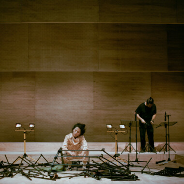 Eine Person in hellem Kostüm sitzt auf einer Bühne zwischen vielen schwarzen Stativen, die im ganzen Raum verteilt sind. Sie scheint etwas mit den Stativen zu bauen. Im Hintergrund steht eine weitere Person, den Kopf nach unten gebeugt.