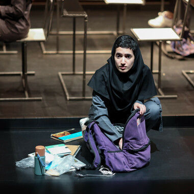In the centre of the image, a person is squatting on the ground. The girl is wearing a black hijab that covers her shoulders but exposes the base of her dark, short hair. The girl reaches with her right hand into the purple backpack on the floor in front of her and has a distressed look on her face. The content of the backpack, consisting of notepads and a bottle, is spread out on the ground to her right. Desks can be seen in the background of the image.