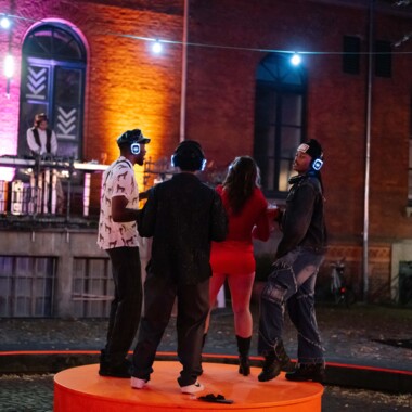 Four people are dancing at a night-time silent disco on an orange-coloured platform under the open sky. They are wearing glowing headphones. In the background, a person stands at a DJ desk on a balcony in front of a colourfully lit brick façade.