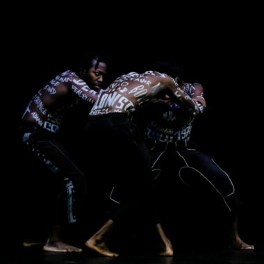 Three PoC whose naked torsos are covered with white lettering. They are barefoot and stand crouched. They touch each other with their arms. The person on the left can be seen from the side and looks intensely at the two other performers who are standing opposite to each other.